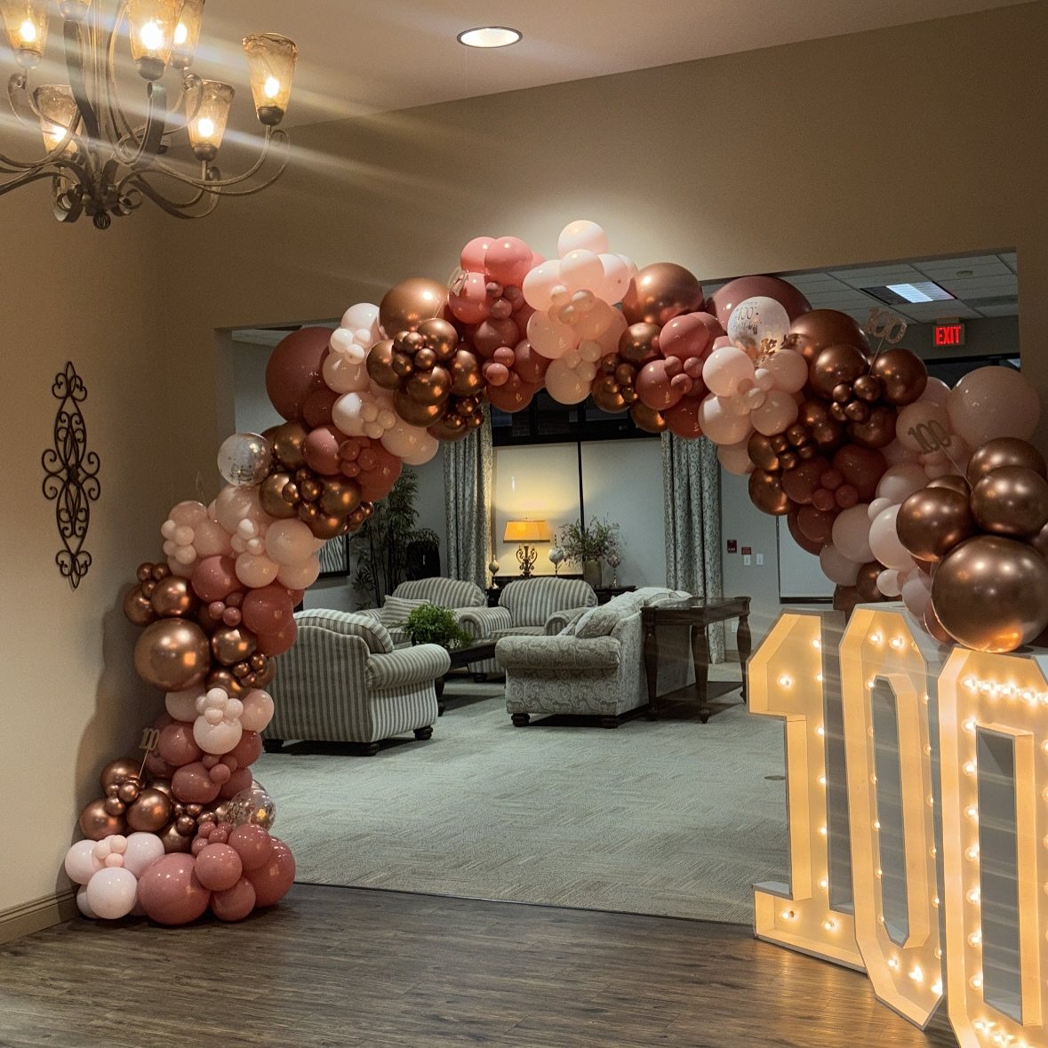 Rose gold balloon arch framing large illuminated marquee numbers reading one hundred.
