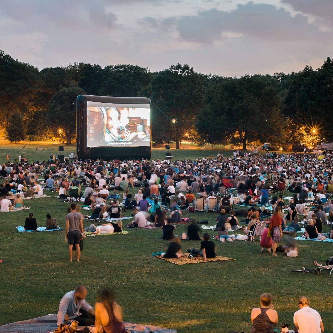 A large crowd sitting on grass watching a movie on a big outdoor screen.