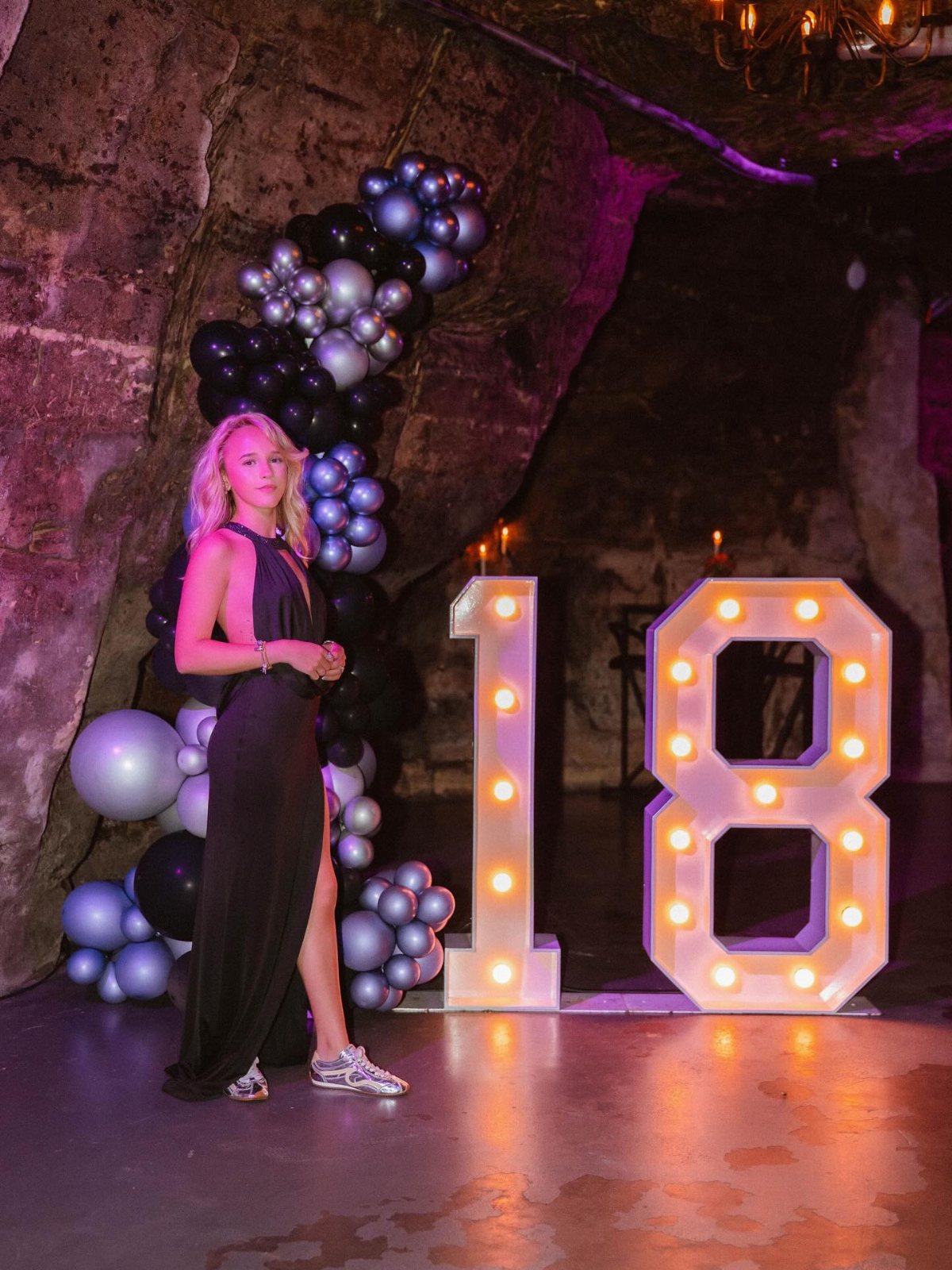 Young woman in a black dress posing next to large glowing number 18 marquee lights.