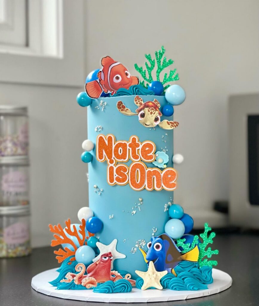 A blue Nemo themed birthday cake with sea creatures and orange text saying Nate is One.