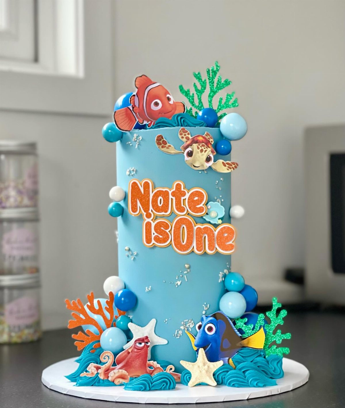 A blue Nemo themed birthday cake with sea creatures and orange text saying Nate is One.