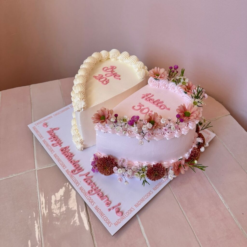 A heart-shaped pink and white birthday cake decorated with small pink and purple flowers.