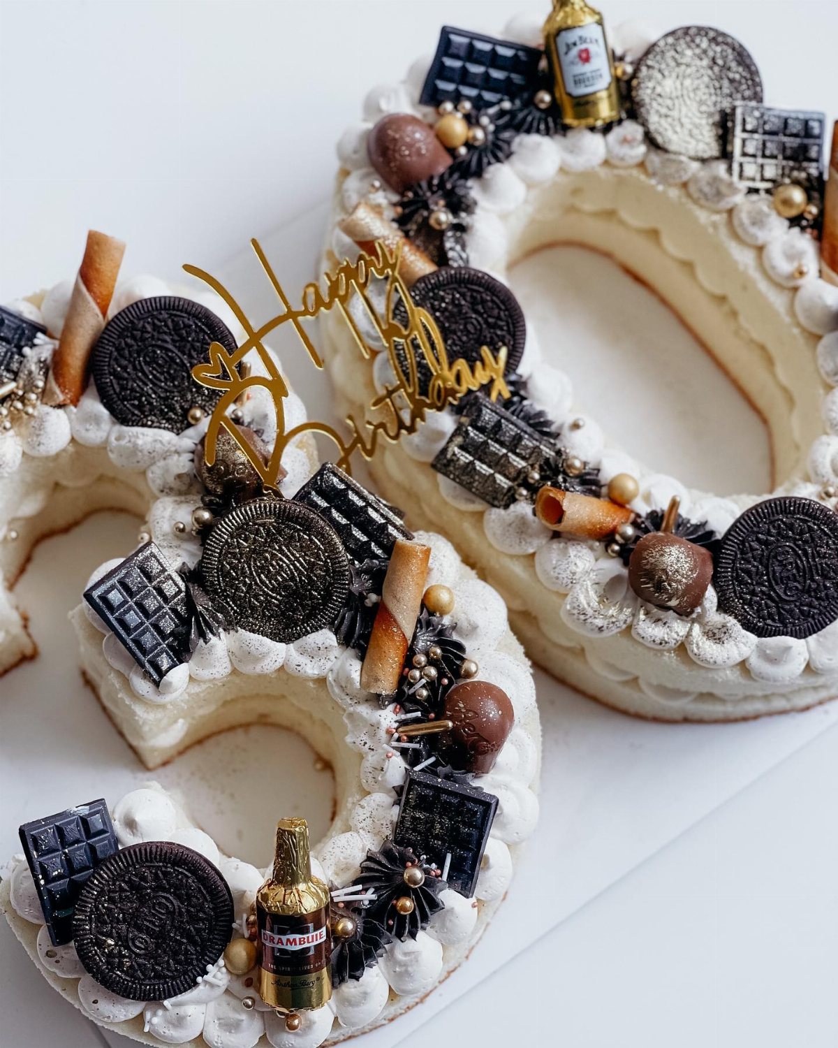 Number thirty shaped cakes decorated with chocolate, cookies, and miniature liquor bottles on a white surface.