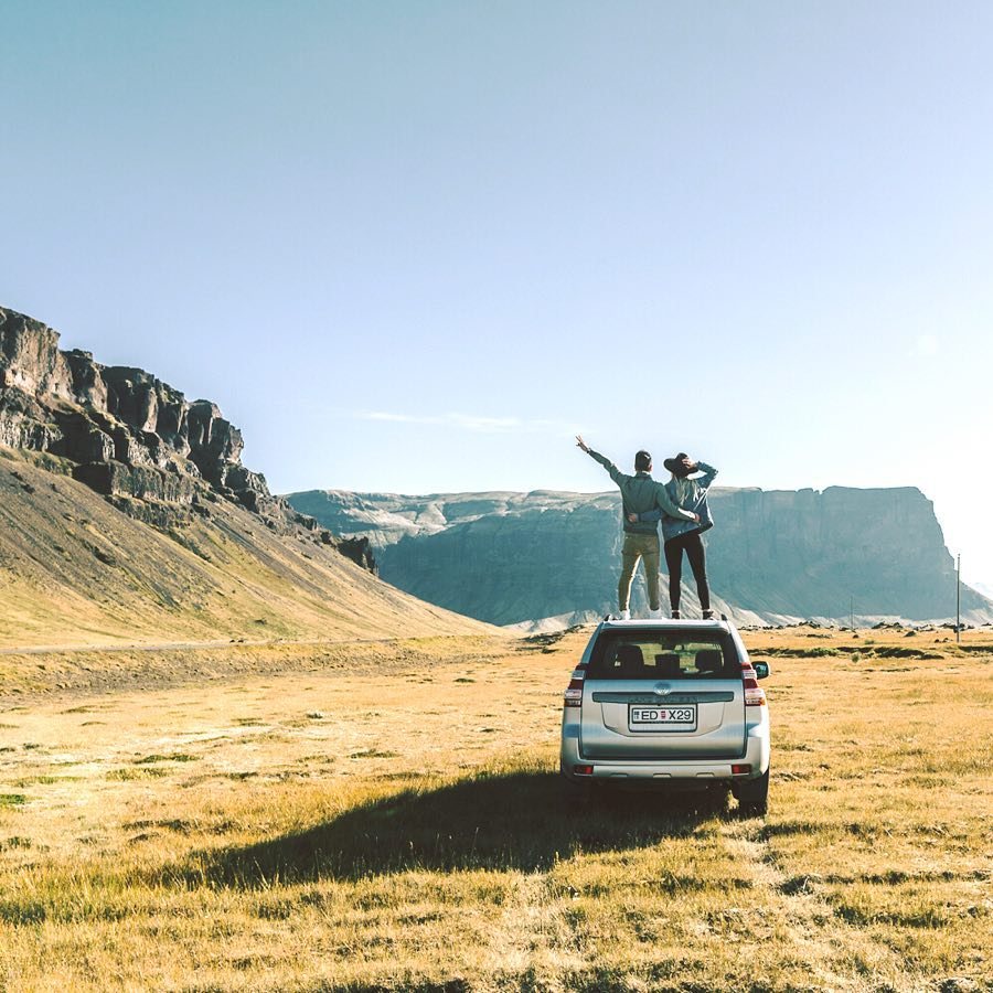 Two people standing on a silver SUV overlooking vast cliffs and grassy plains.