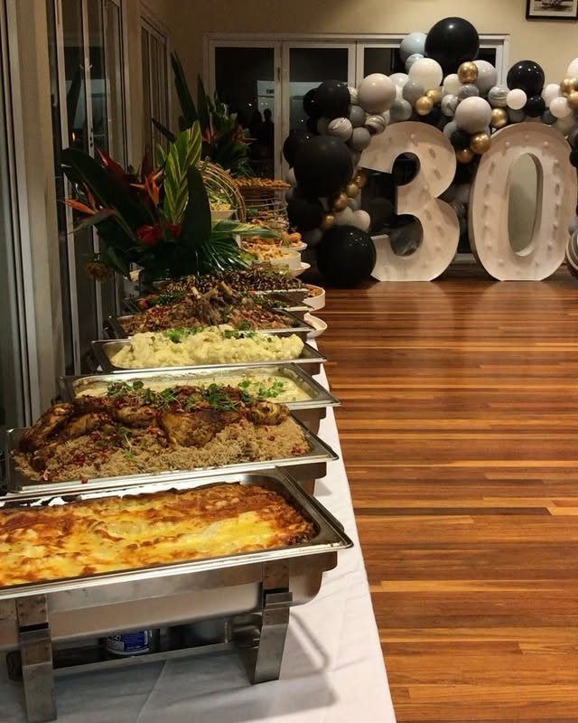 Buffet table with various hot dishes in silver chafing dishes and large number thirty decorations.