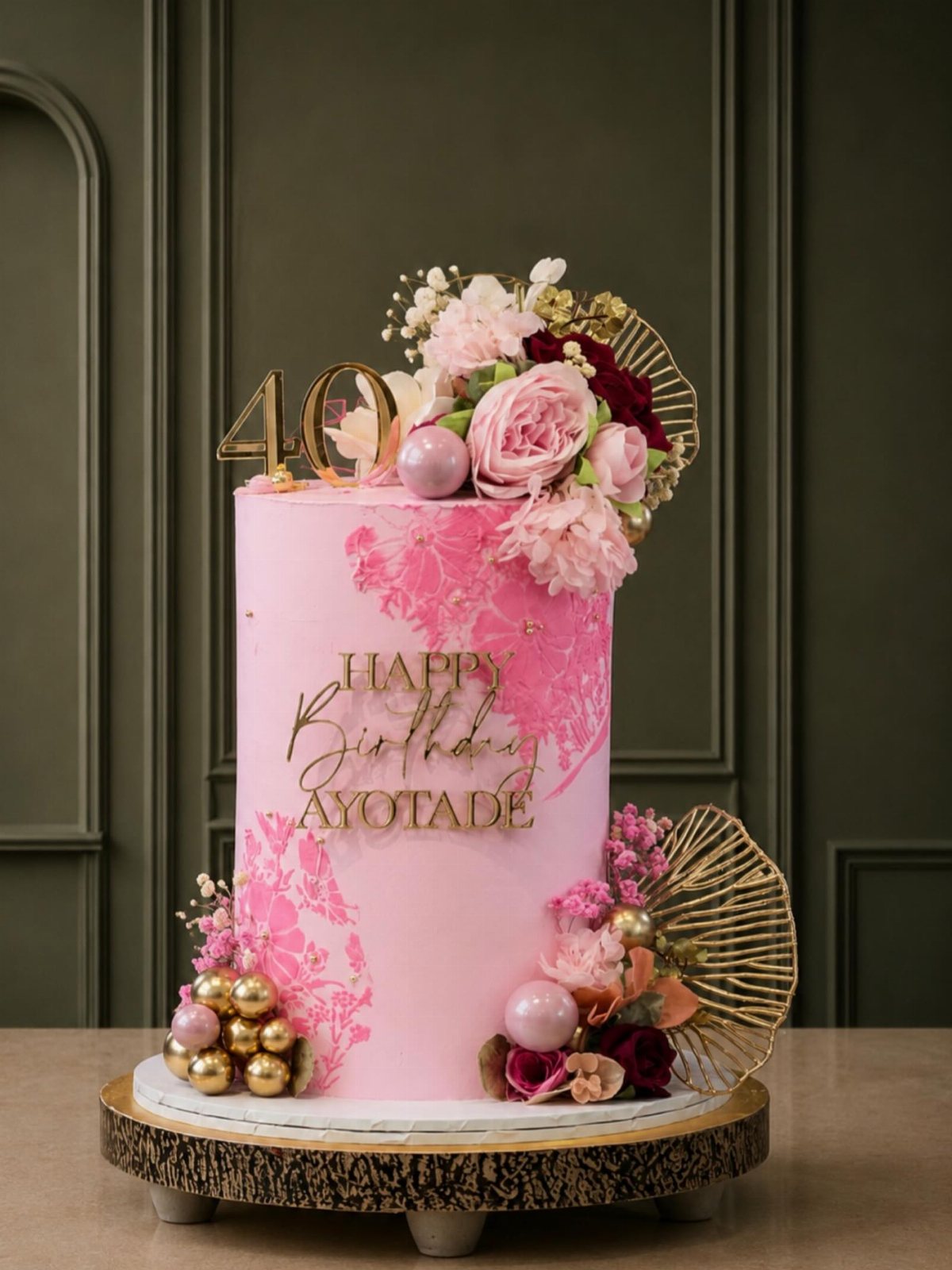 Pink birthday cake with gold number 40, pink flowers, and gold decorative accents on a wooden stand.