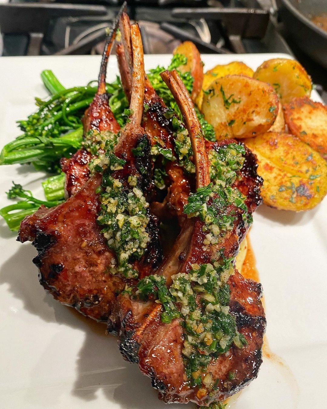 Grilled lamb chops with herb sauce, roasted potatoes, and green vegetables on a white plate.