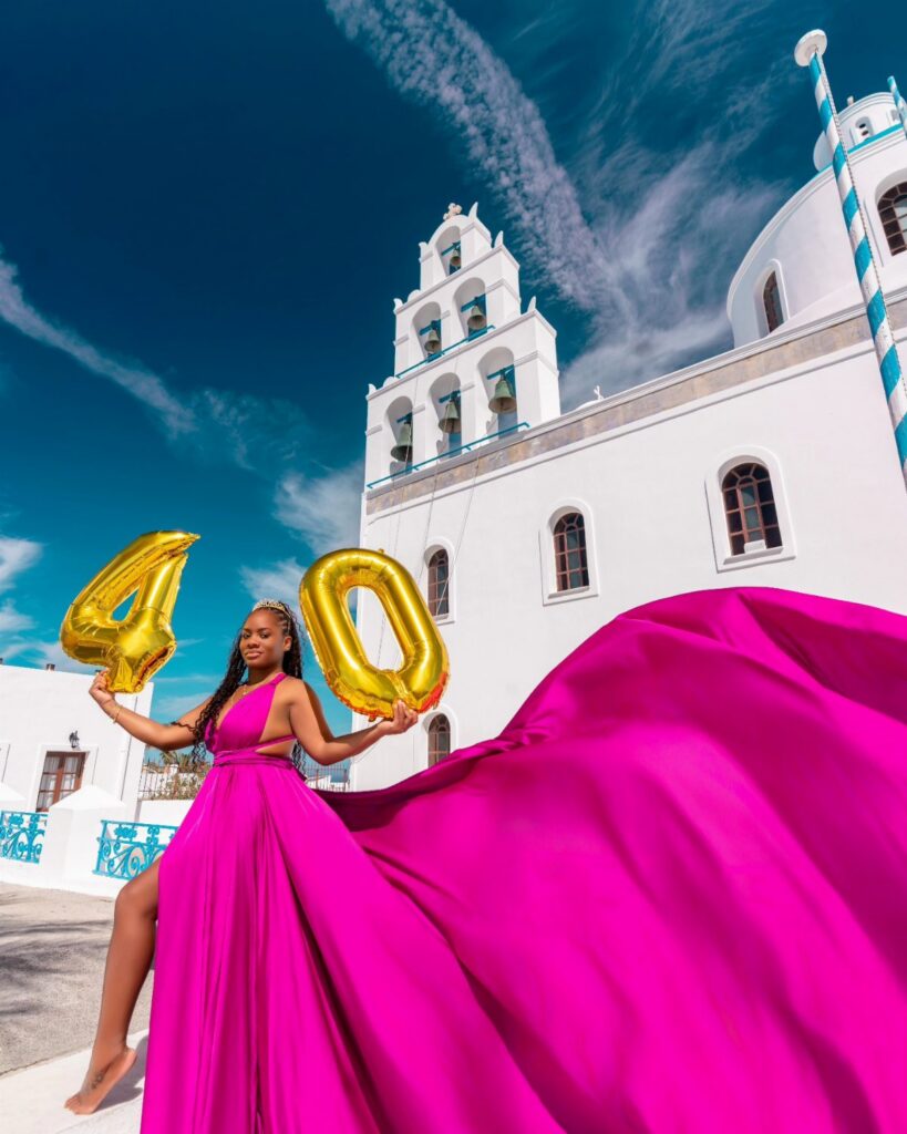 Woman in a magenta gown holding gold number 40 balloons in front of a white church.