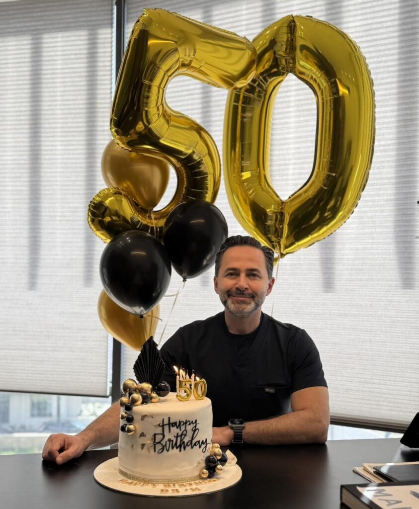 A smiling man sitting behind a birthday cake with large gold number fifty balloons.