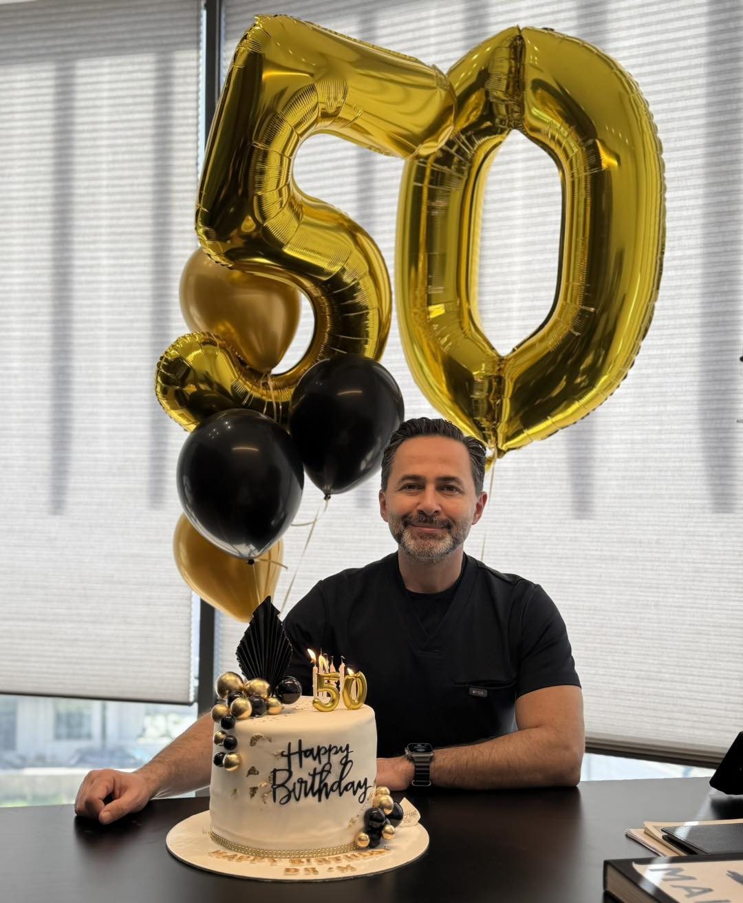 A smiling man sitting behind a birthday cake with large gold number fifty balloons.