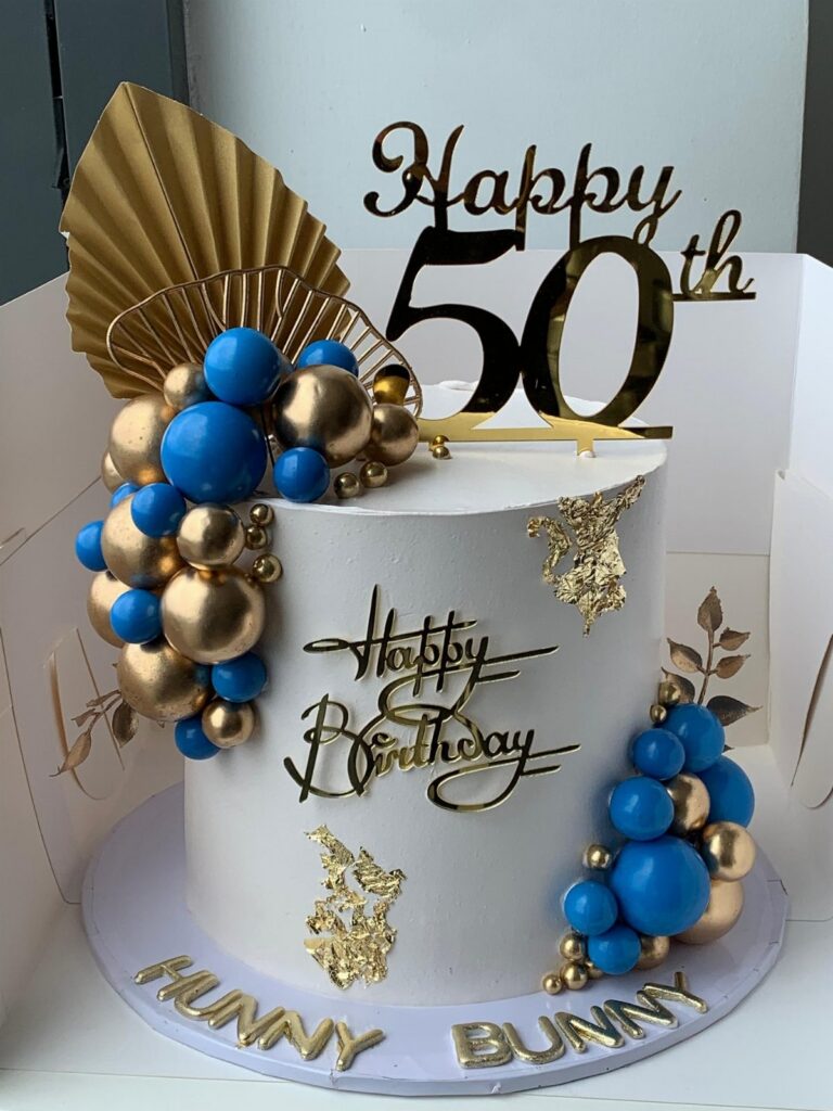 White birthday cake with gold and blue sphere decorations and a Happy 50th topper.
