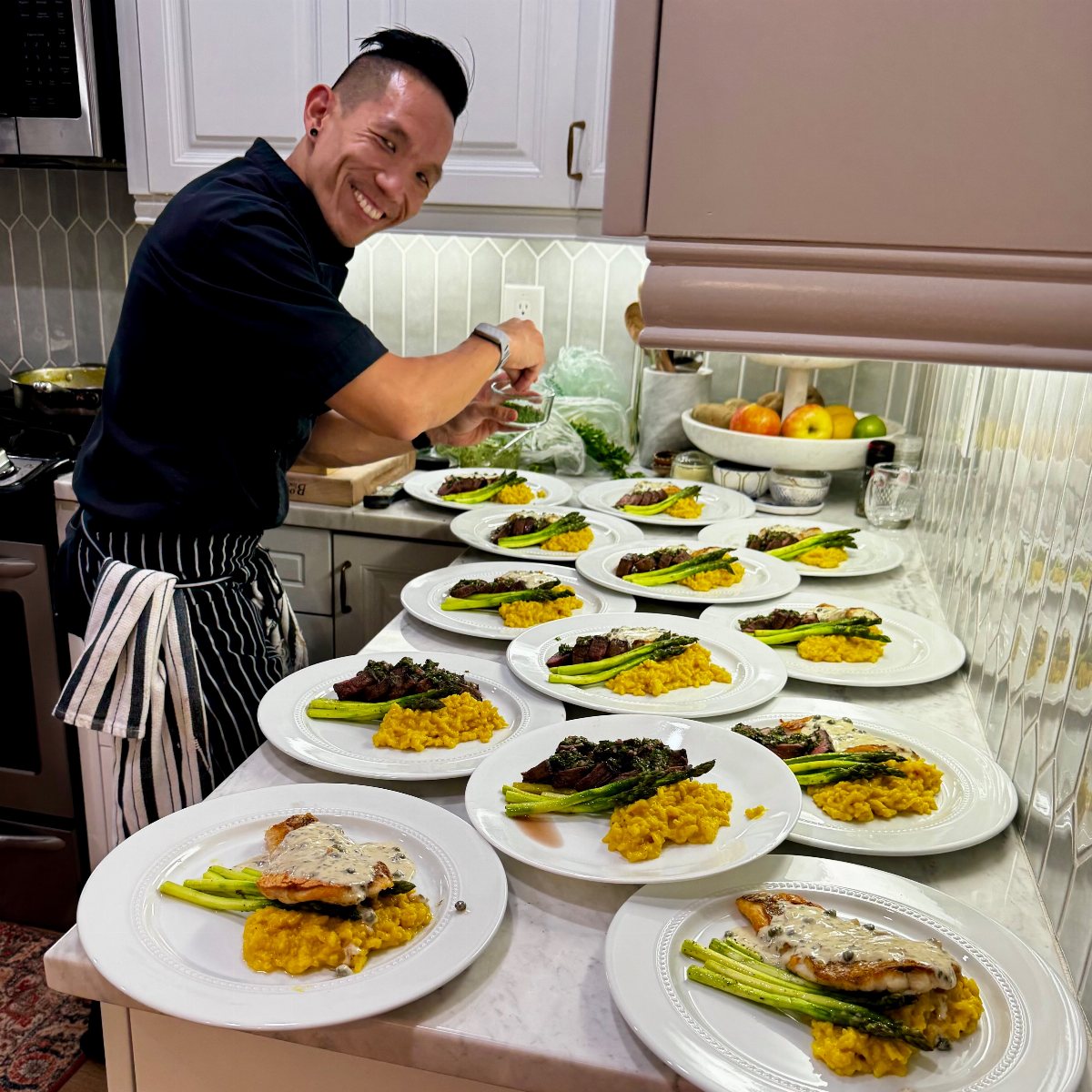 A smiling chef prepares several plates of fish, asparagus, and risotto in a bright kitchen.