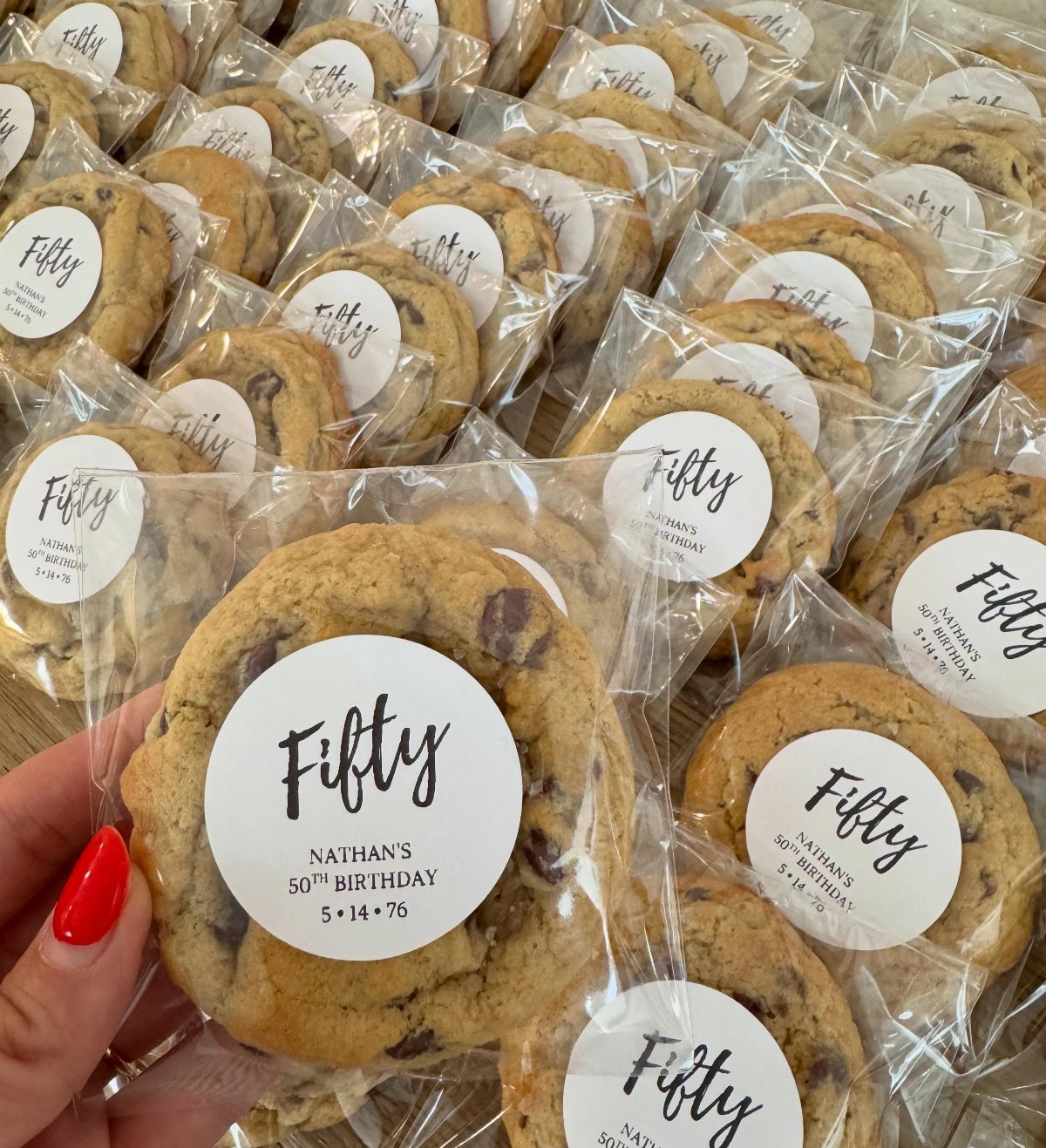 A hand holding a wrapped chocolate chip cookie with a custom Fifty birthday sticker.