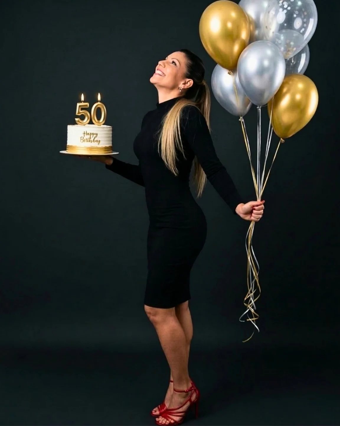 Smiling woman in a black dress holding a birthday cake with fifty candles and gold balloons.
