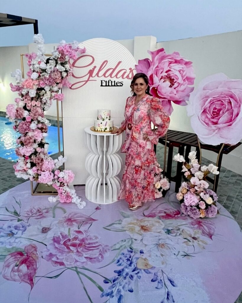 Woman in a pink floral dress standing by a decorated cake pedestal and floral backdrop.