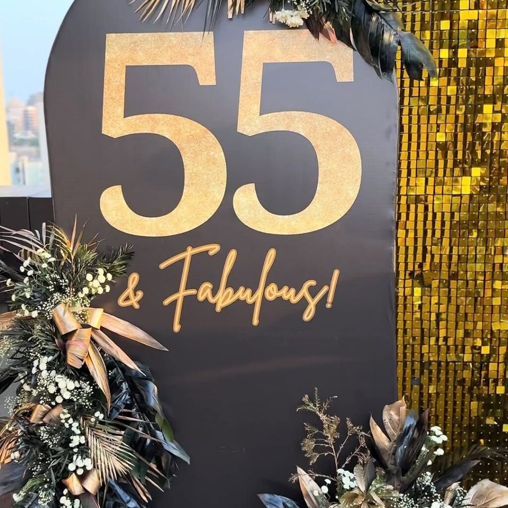 A black sign with gold text reading 55 & Fabulous surrounded by dark floral arrangements.