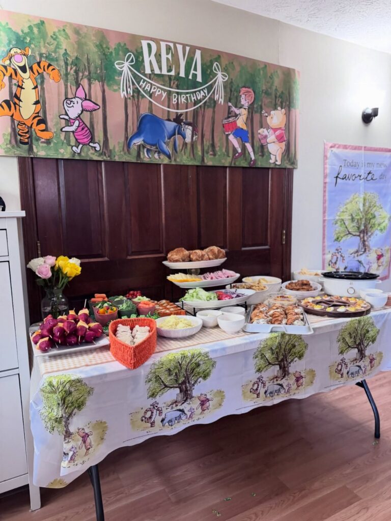 A buffet table filled with party food under a Winnie the Pooh themed birthday banner.
