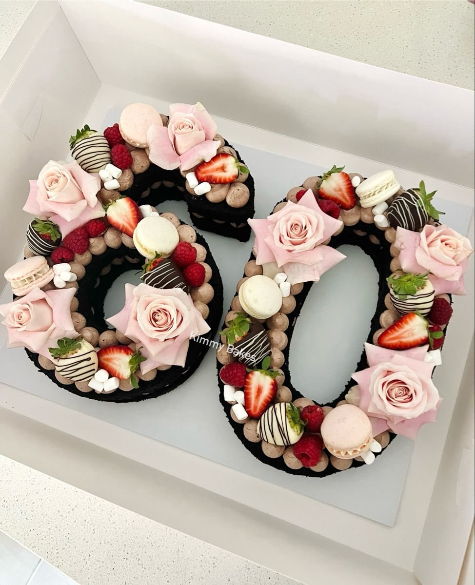 Number sixty cake decorated with pink roses, macarons, and fresh berries in a white box.