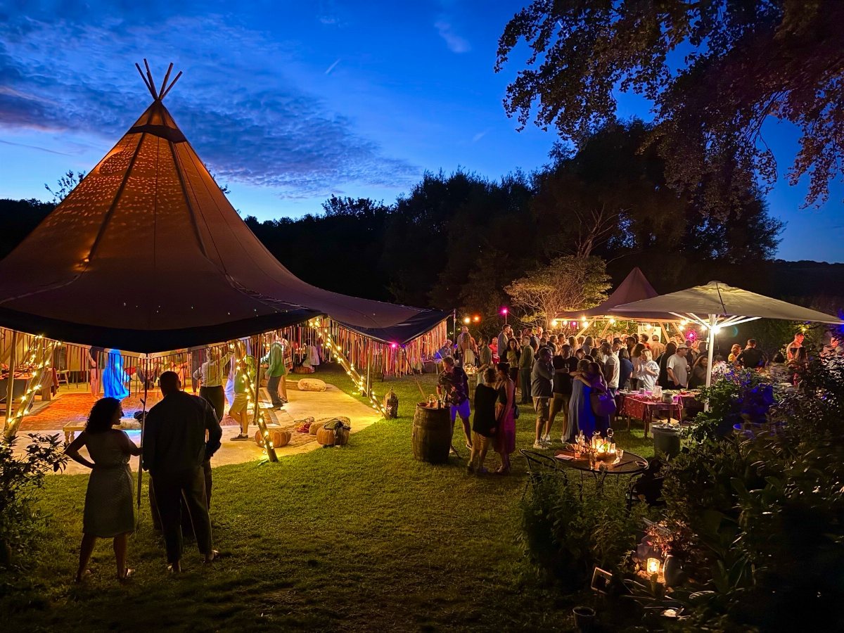 People socializing at an outdoor evening party under a large illuminated tipi tent.