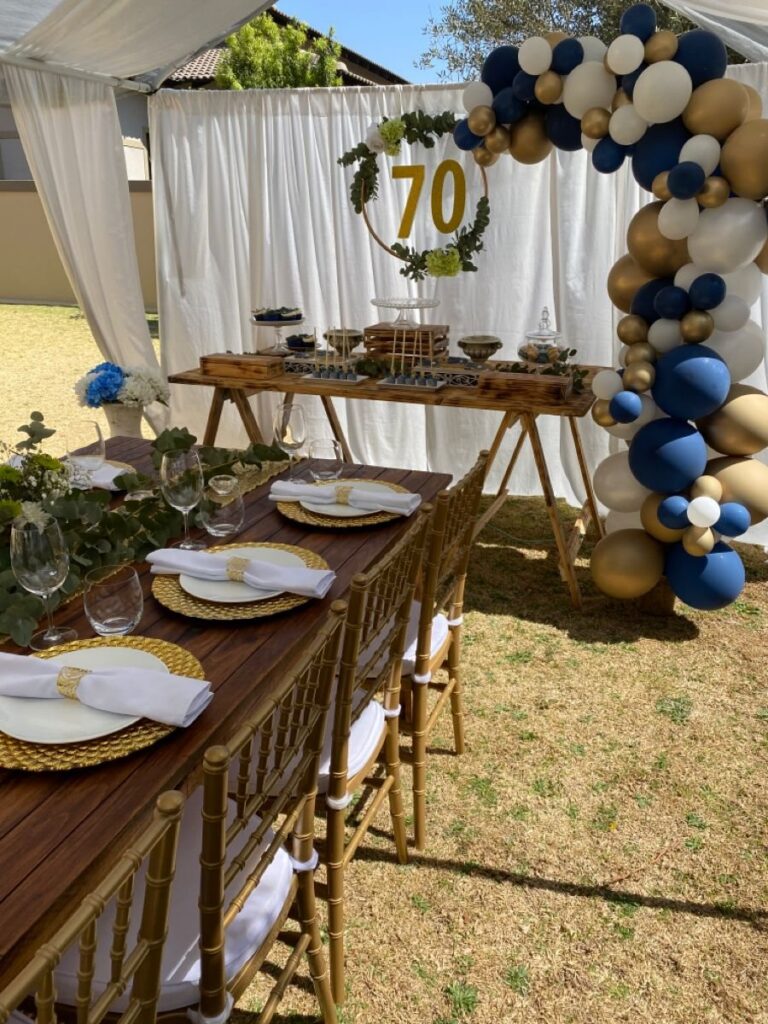 A decorated outdoor dining table with gold accents and a balloon arch for a 70th birthday.