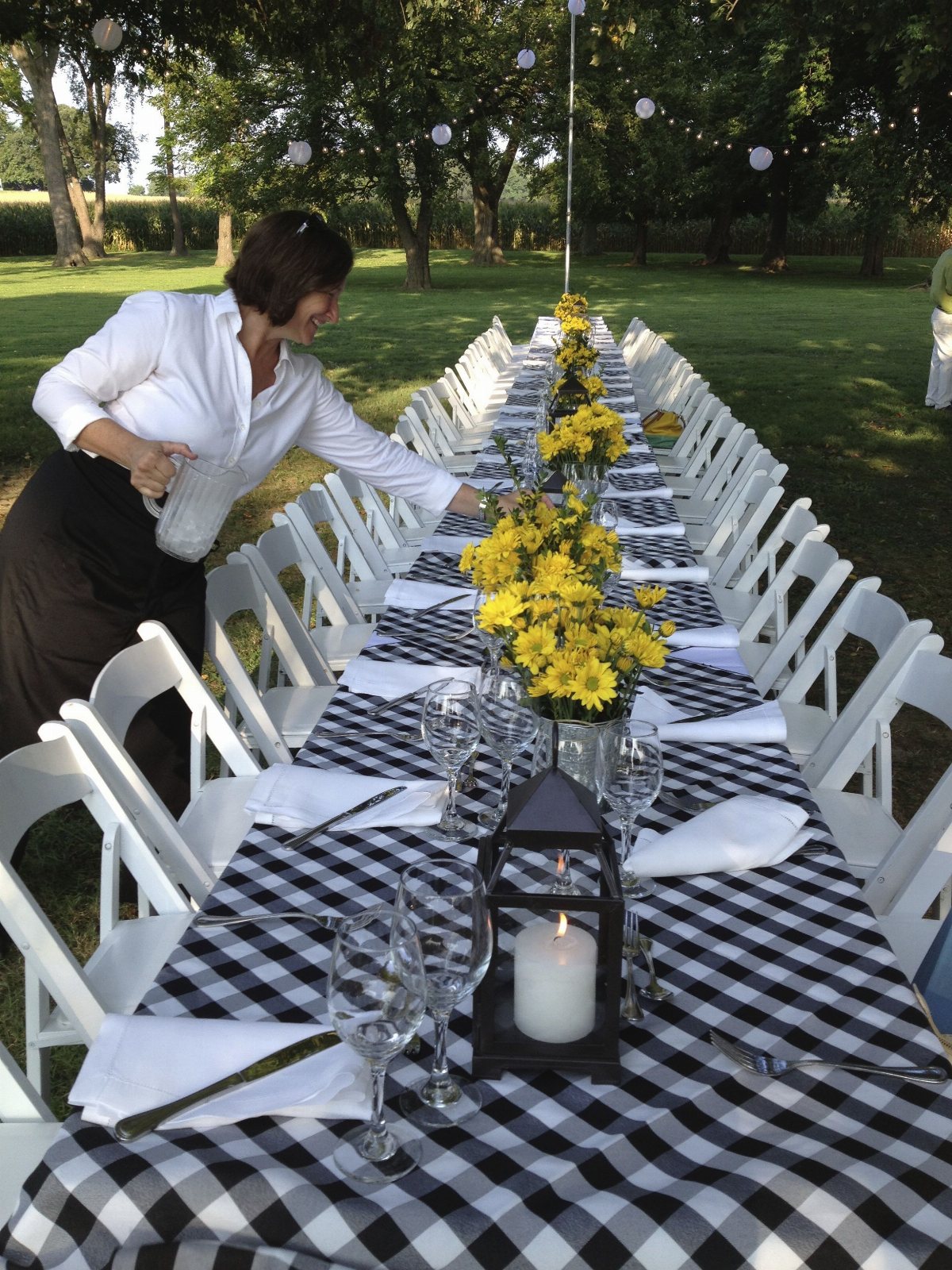 A woman setting a long outdoor dining table with checkered tablecloth and yellow flowers.