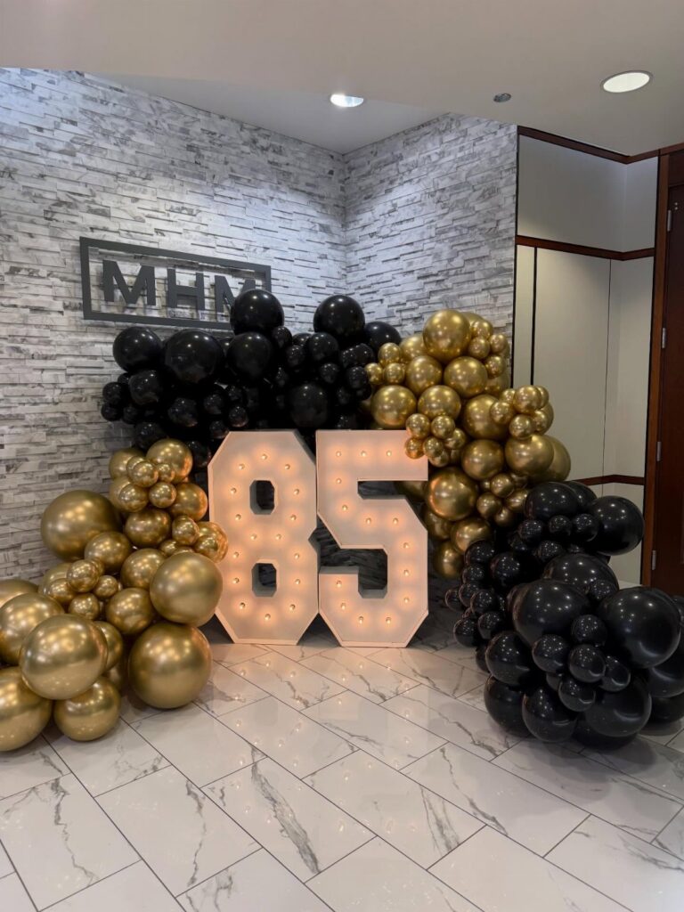 Large illuminated marquee numbers 85 surrounded by black and gold balloon arrangements on a tiled floor.