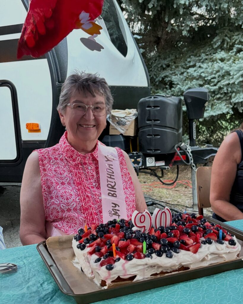 Smiling elderly woman wearing a birthday sash sitting behind a large berry sheet cake.