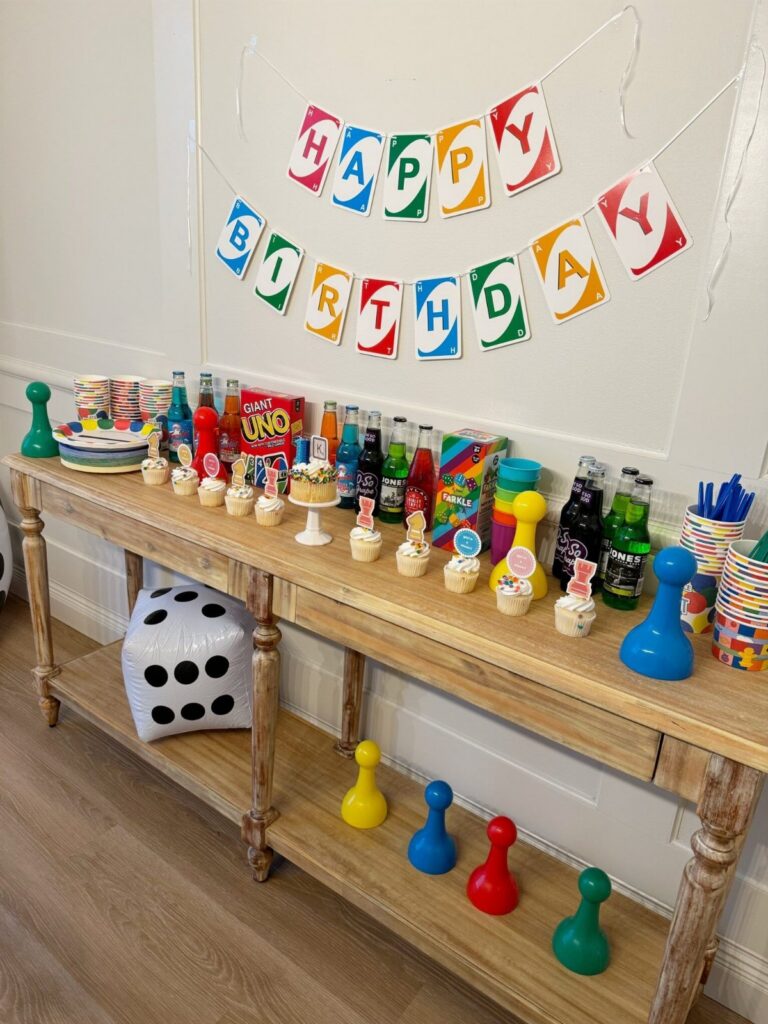 A wooden table decorated with colorful games, sodas, cupcakes, and bowling pins for a party.
