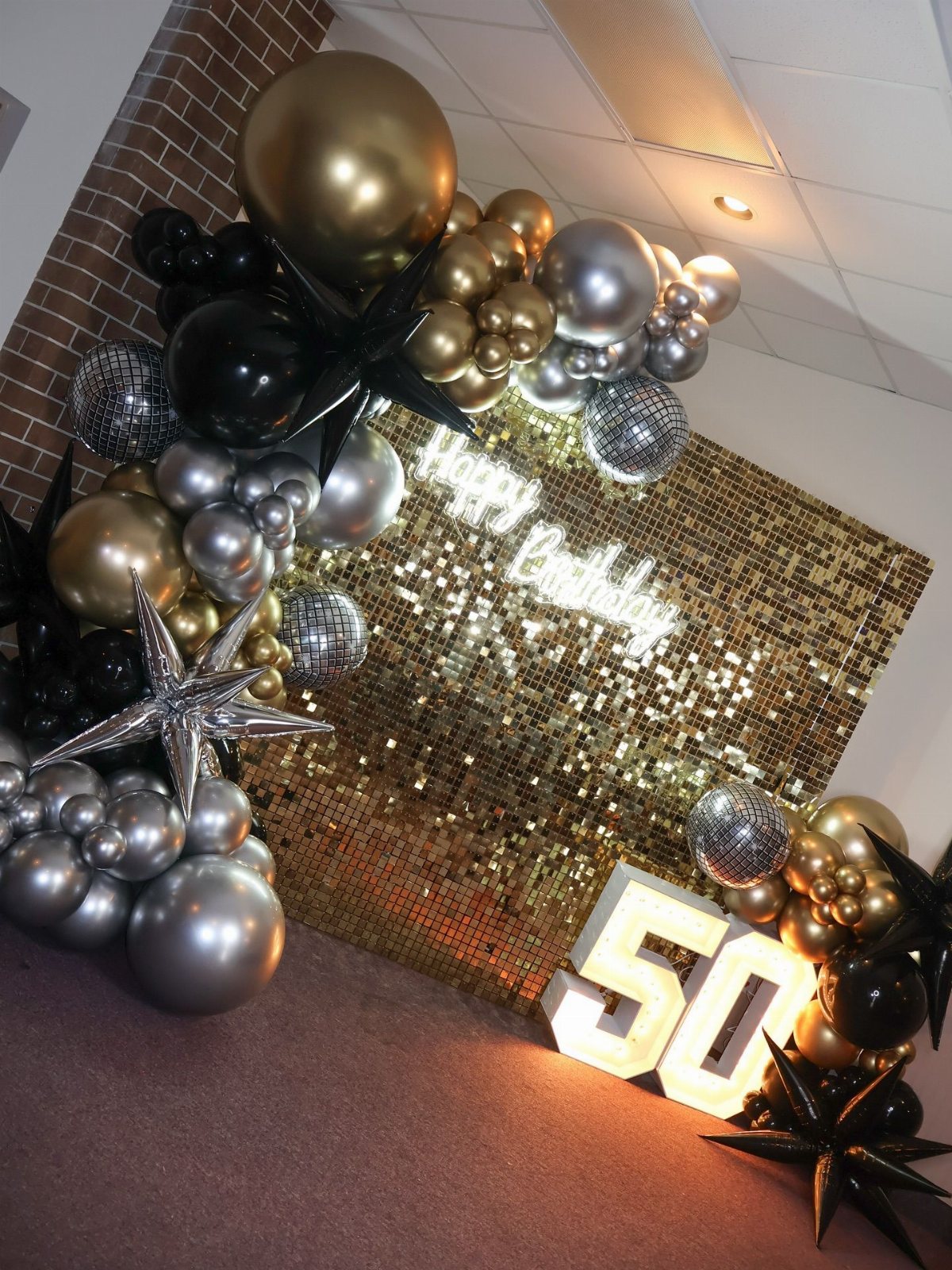 Gold sequin backdrop with black and silver balloons and large illuminated number 50.