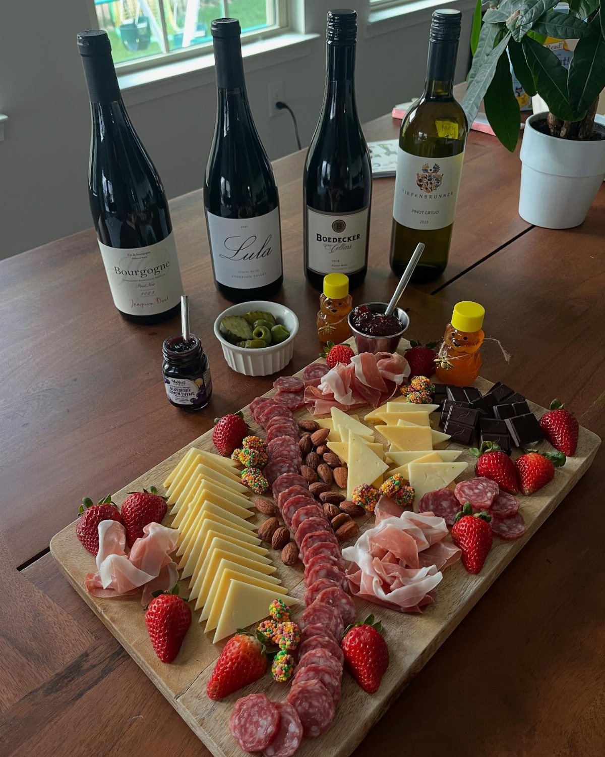 A wooden charcuterie board with meats, cheeses, and strawberries next to four wine bottles.