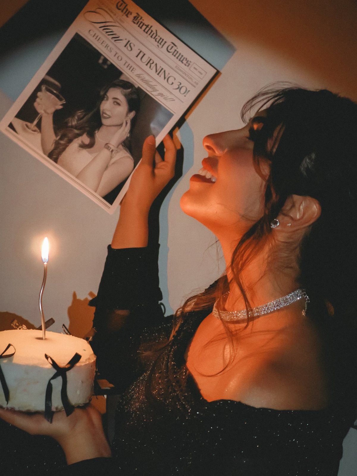 Woman in a black dress holding a birthday newspaper and a small lit candle cake.