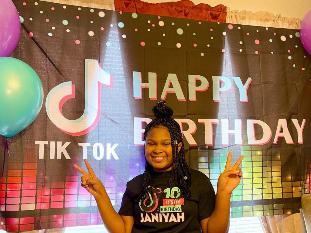 A smiling girl posing in front of a TikTok themed happy birthday backdrop.