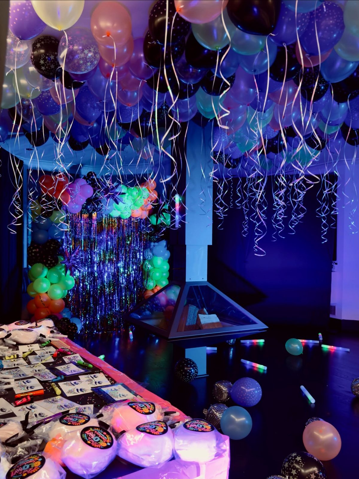 Colorful balloons with silver streamers hang from the ceiling above a table of party favors.
