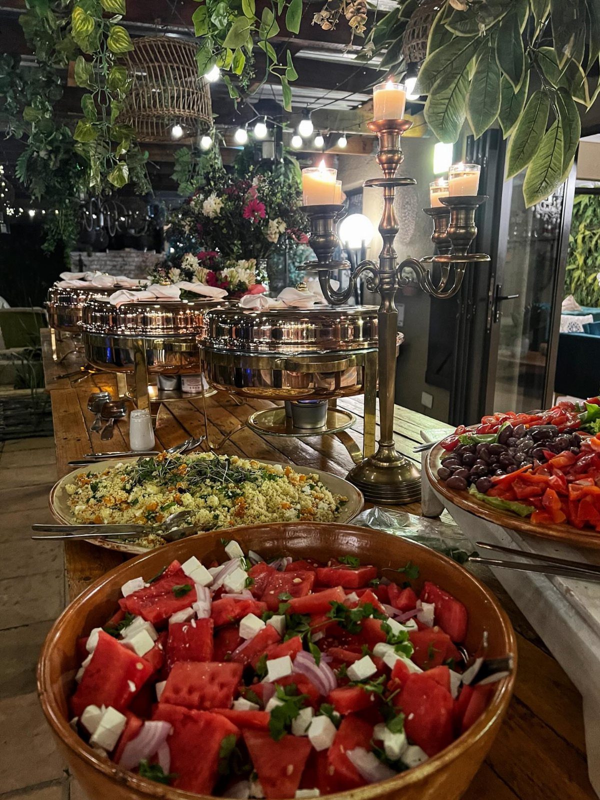 A buffet table featuring watermelon salad, rice, and copper chafing dishes under warm candlelight.