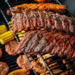 Grilled ribs, chicken wings, sausages, and corn on the cob cooking on a black barbecue grate.