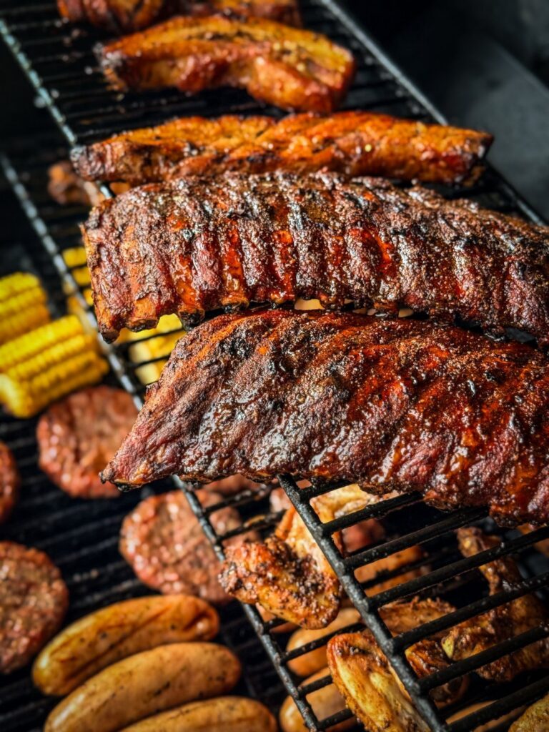 Grilled ribs, chicken wings, sausages, and corn on the cob cooking on a black barbecue grate.