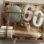 A wooden gift box containing a teal heart, white number sixty, and a happy birthday candle.