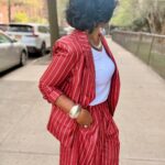 A woman in a red pinstriped suit standing on a city sidewalk wearing gold jewelry.