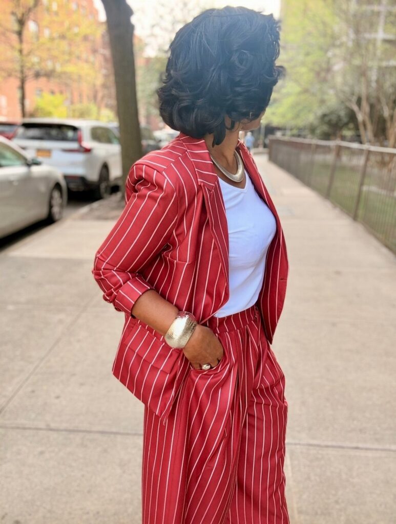 A woman in a red pinstriped suit standing on a city sidewalk wearing gold jewelry.