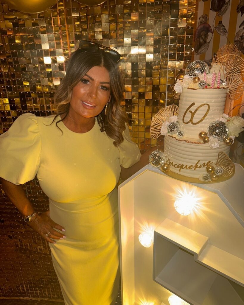 A woman in a yellow dress standing next to a decorated 60th birthday cake.